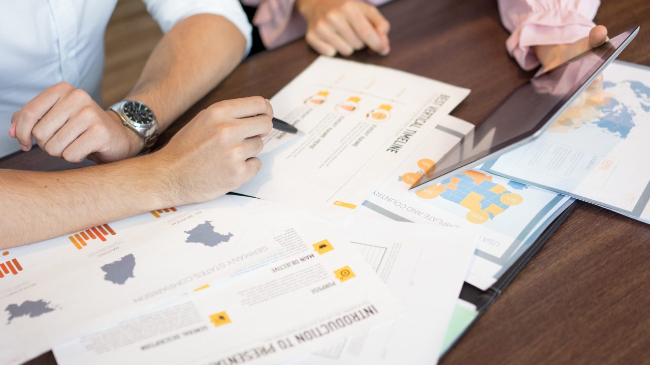 Hands of businesspeople discussing marketing reports and presentation on tablet. Close-up of young Caucasian businessman and businesswoman sitting at table and working over documents. Teamwork concept